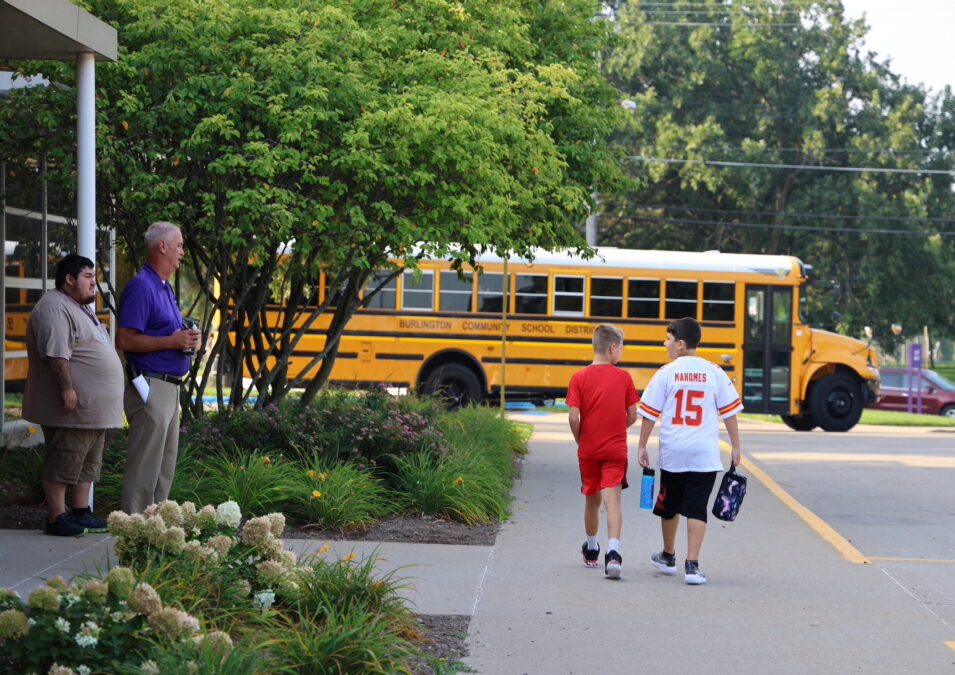 Two elementary students walk alongside a school toward a school bus