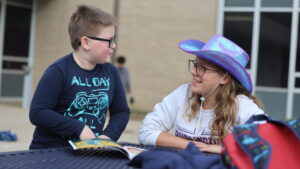 A high school student wears a cowboy hat for Hat Day while reading a book with her elementary Club M mentee outside the cafeteria of Burlington High School