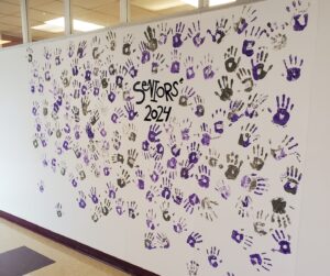 Handprints made with grey and purple paint are shown on a wall around the words "Seniors 2024"