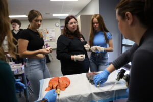 Three high school girls stand behind a table that holds two sets of lungs, one healthy and the other diseased, as their teacher and a guest speaker talk about the lungs.