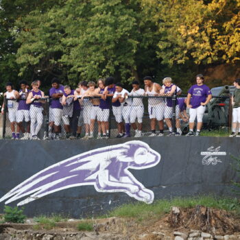 Members of the varsity football team stand on top of a wall displaying the Grayhound logo while watching the JV team play below.