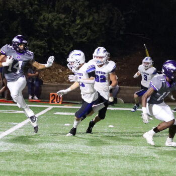 A football player cradles the ball in one arm while holding up the other to fend off members of the opposing team.