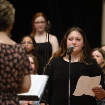 A student holds a piece of paper while standing in front of a microphone during a choir concert.