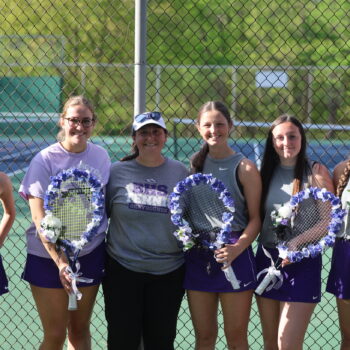 The girls tennis coach stands alongside senior team members while posing for a photo on senior night. The players are holding tennis rackets wreathed by purple flowers.