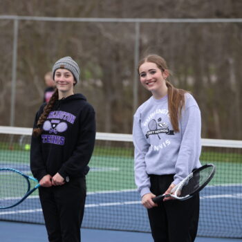 Two members of the BHS girls tennis team smile for a photo at the start of their doubles match. It's early in the season, so they're dressed for cool weather.