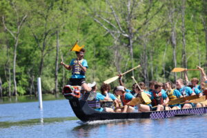 A dragon boat glides over water as its student passengers cheer on and the flag catcher holds up the flag.