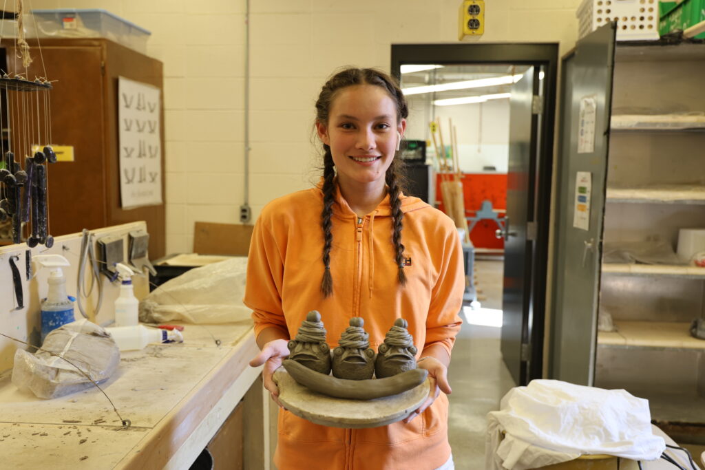 A high school student smiles while holding up a clay sculpture she created