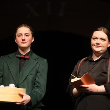 A student actor holds a box full of glowing green bottles while another dressed as a reporter takes notes during the spring play, 