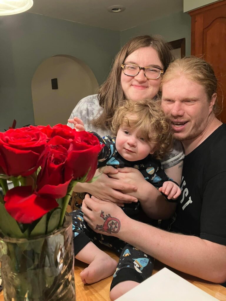 Allison Cook poses for a photo with her son and fiance next to a bouquet of roses.