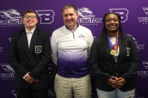 Two students wearing DECA blazers and medals stand on either side of their club advisor with a BCSD banner in the background