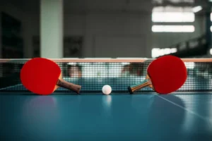 Two table tennis paddles rest on a ping pong table with a ball in the middle and a net in the background