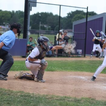 Dust flies from a catchers mit as it closes around a baseball while a member of the opposing team stands at home base holding a bat.