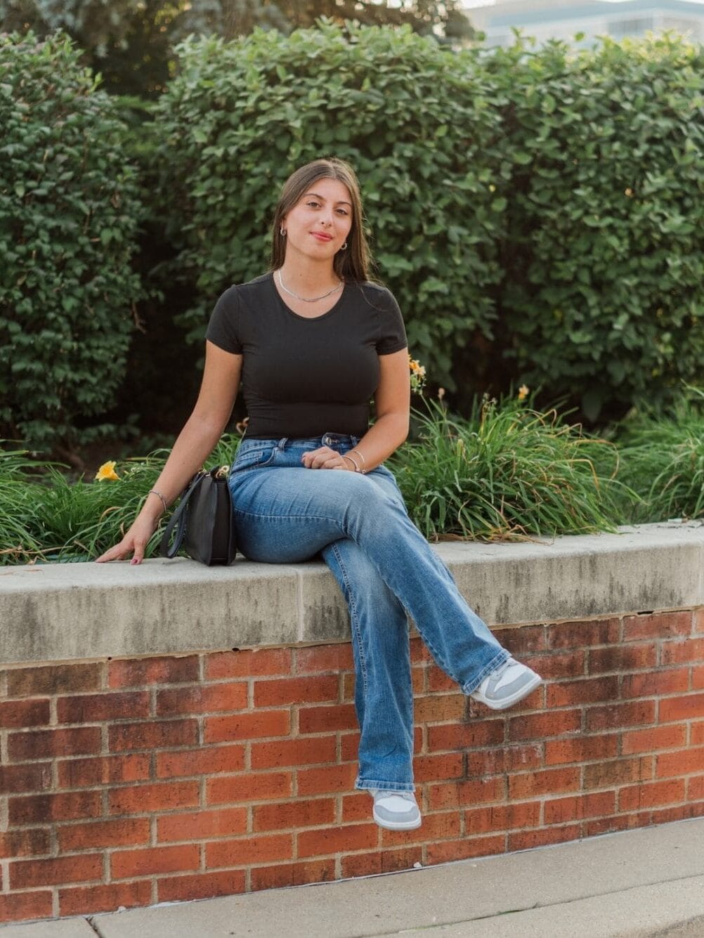 Rebecca Imbimbo poses for a photo while sitting on a brick retaining wall with bushes and plants in the background.