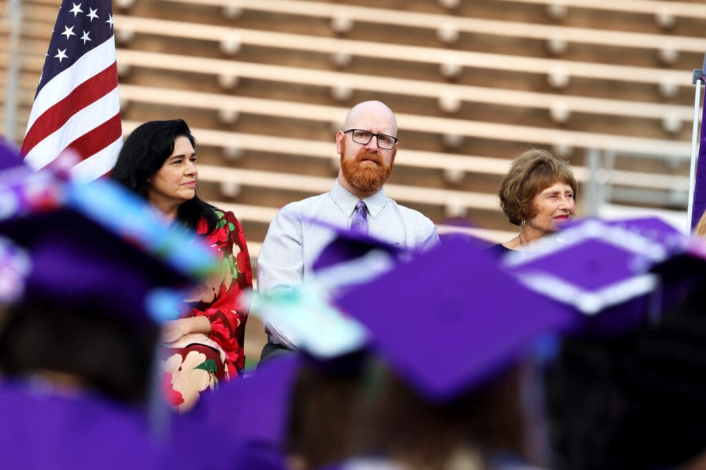 Human Resources Director Laci Johnson and Burlington School Board members William Vance and Deborah Hatteburg are seated behind a sea of purple caps during Burlington High School's 2025 commencement ceremony at Bracewell Stadium.
