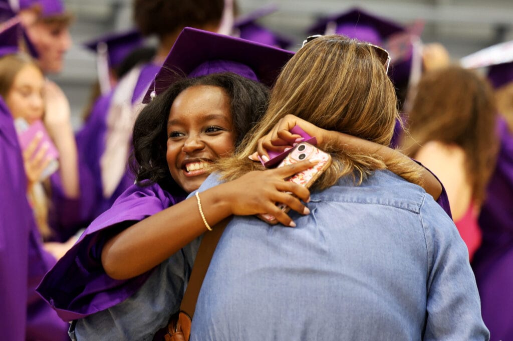 A graduating senior wearing a purple cap and gown smiles while hugging a teacher prior to the start of Burlington High School's commencement ceremony Saturday, May 31, 2025, inside Clark Field House.