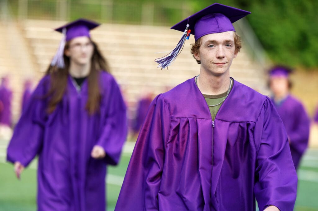 A graduating senior wearing a cap and gown smiles while making his way to his seat at the start of Burlington High School's 2025 commencement ceremony at Bracewell Stadium.