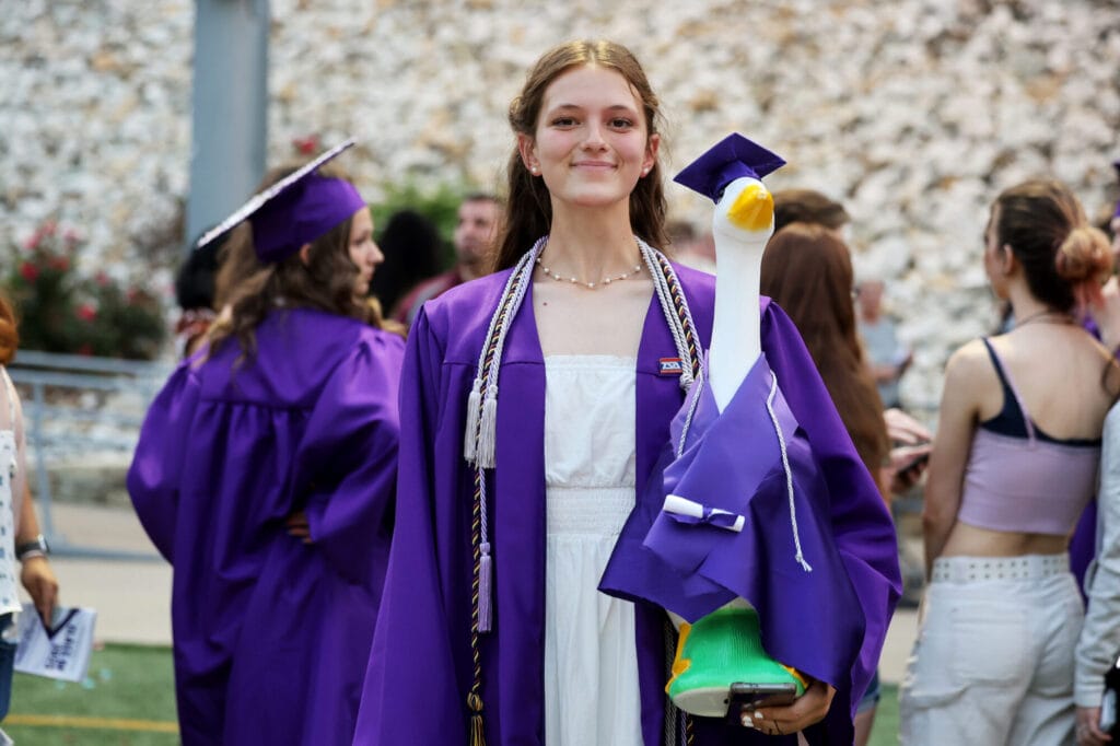 A graduate wearing a purple gown smiles and holds a silly goose dressed to match (also in a purple gown) following Burlington High School's 2025 commencement ceremony at Bracewell Stadium.