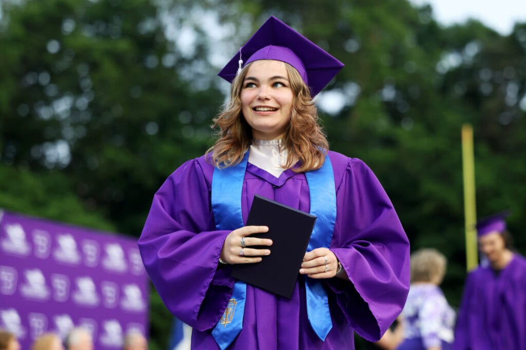A graduate wearing a purple cap and gown smiles while holding up her diploma during Burlington High School's 2025 commencement ceremony at Bracewell Stadium.