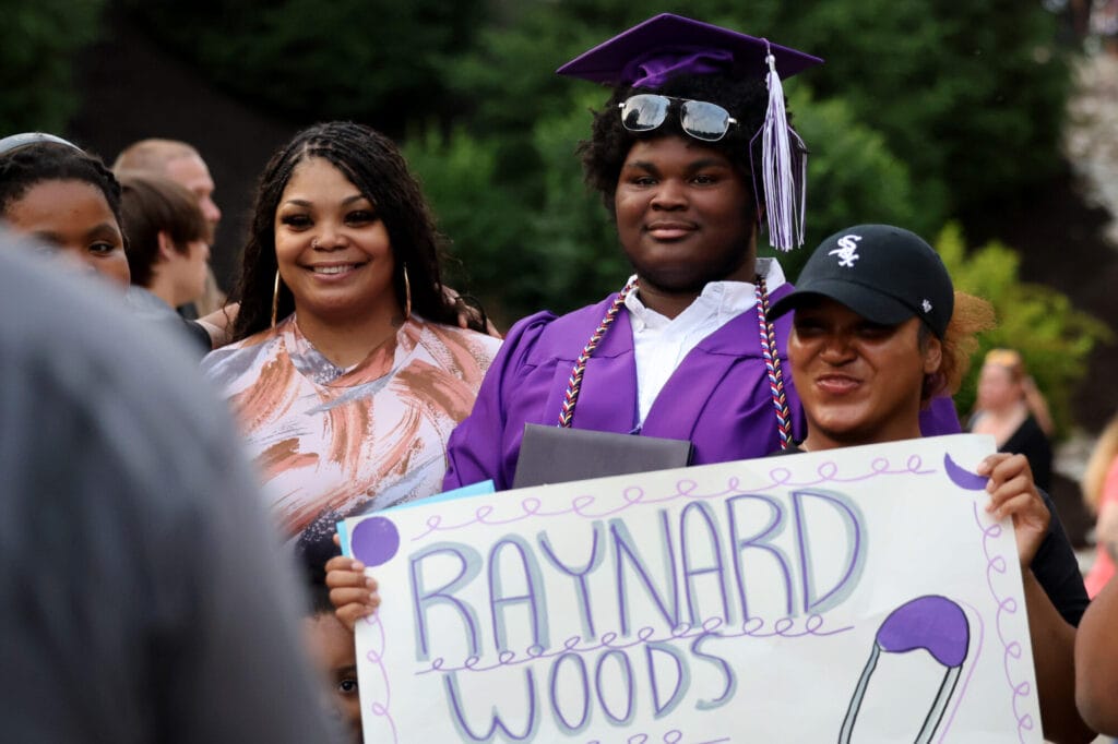 A graduate flanked on either side by family members holding a sign bearing his name, "Raynard Woods," wears a confident yet subtle smile while posing for a photo following Burlington High School's 2025 commencement ceremony at Bracewell Stadium.