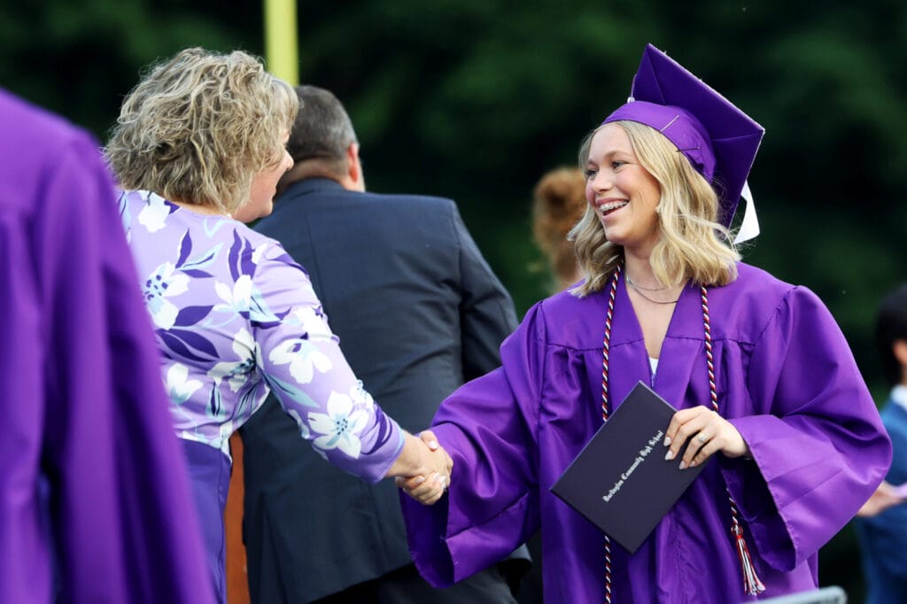 A student wearing a purple cap and gown smiles while shaking hands with the school board president after accepting her diploma during Burlington High School's 2025 commencement ceremony at Bracewell Stadium.