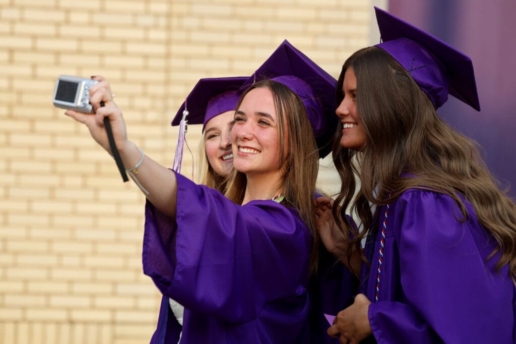 Three graduating seniors smile while posing for a selfie as the one in the middle holds up a camera (not a cellphone) ahead of Burlington High School's 2025 commencement ceremony at Bracewell Stadium.