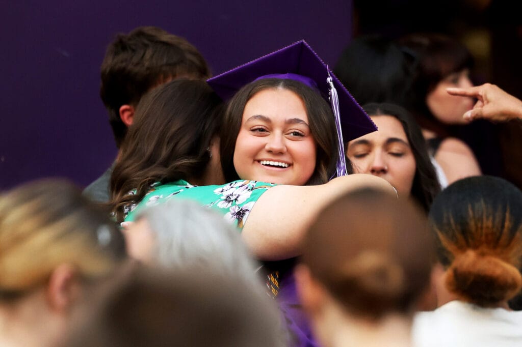 A graduate wearing a purple cap and gown smiles while embracing a family member following Burlington High School's 2025 commencement ceremony at Bracewell Stadium.