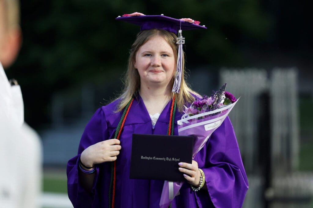 A graduate wearing a purple cap and gown smiles while holding her diploma and a bouquet of purple flowers following Burlington High School's 2025 commencement ceremony at Bracewell Stadium.