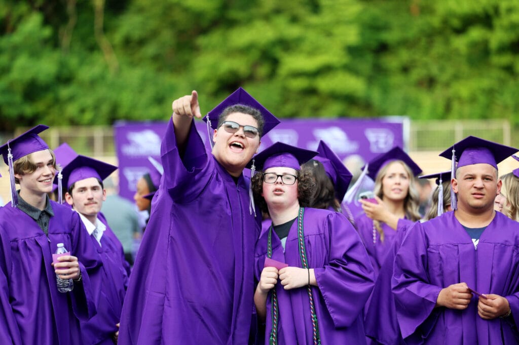A graduating senior smiles and points to someone in the crowd while standing alongside his friends at Bracewell Stadium during Burlington High School's 2025 commencement ceremony.