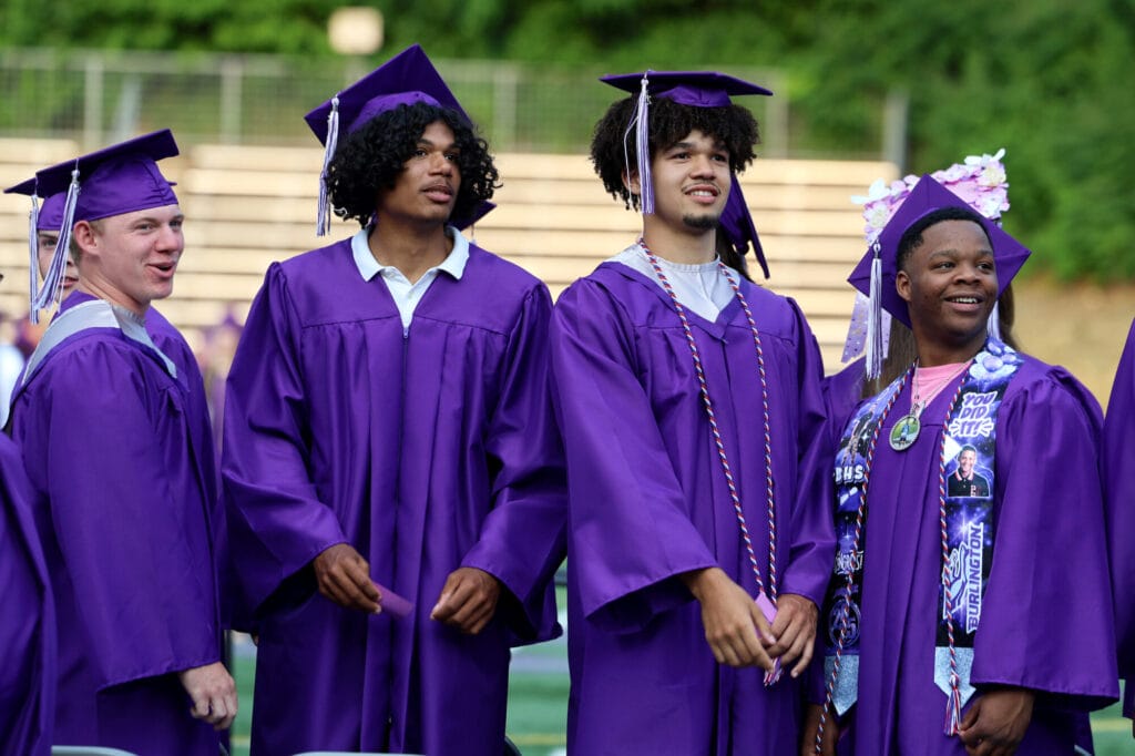 Four graduating seniors wearing caps and gowns look toward the crowd before taking their seats during Burlington High School's 2025 commencement ceremony at Bracewell Stadium.
