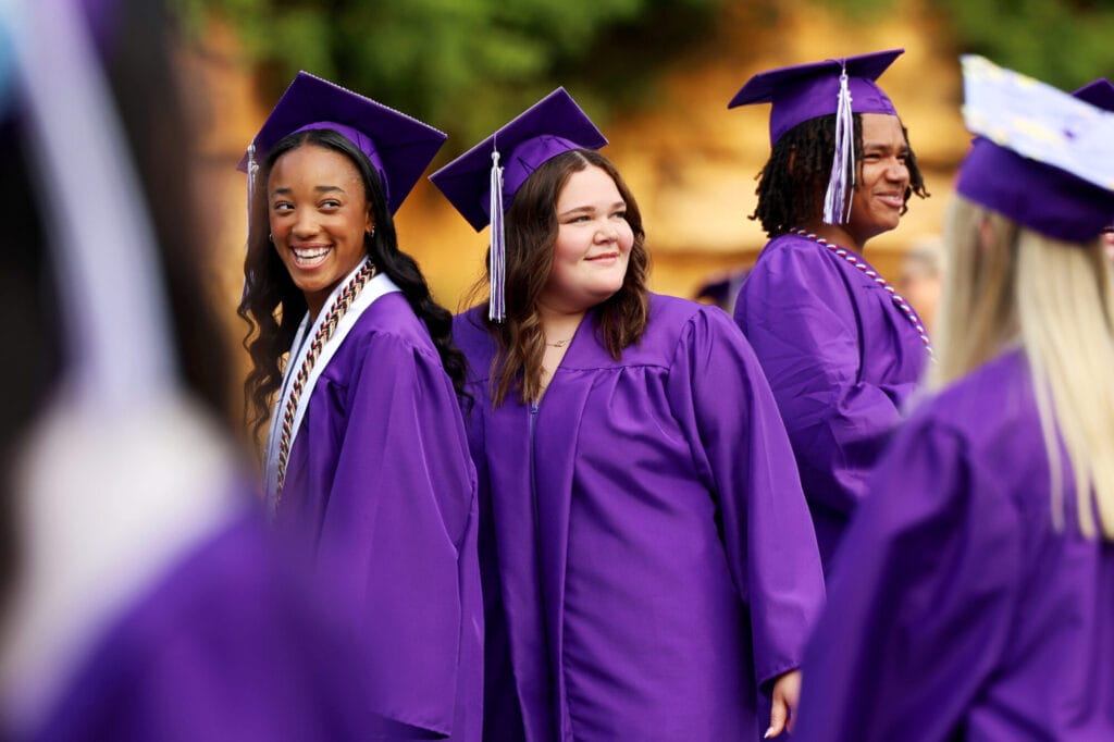 Three graduating seniors wearing purple caps and gowns smile while facing the crowd before taking their seats during Burlington High School's 2025 commencement ceremony at Bracewell Stadium.