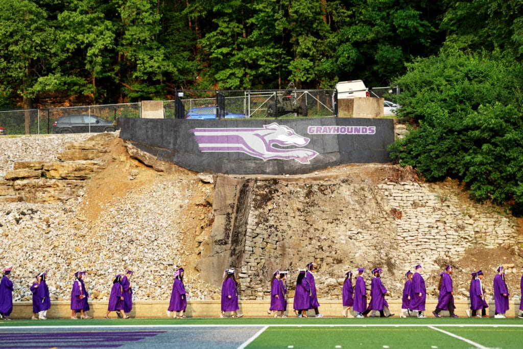 Graduating seniors wearing purple caps and gowns walk beneath on Grayhound mural while en route to their seats at the start of Burlington High School's 2025.