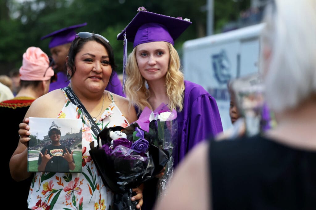 The mother of Zephy Johnson holds up her daughter's photo and a bouquet of purple flowers while posing for a photo alongside her daughter's friend following Burlington High School's 2025 commencement ceremony at Bracewell Stadium.