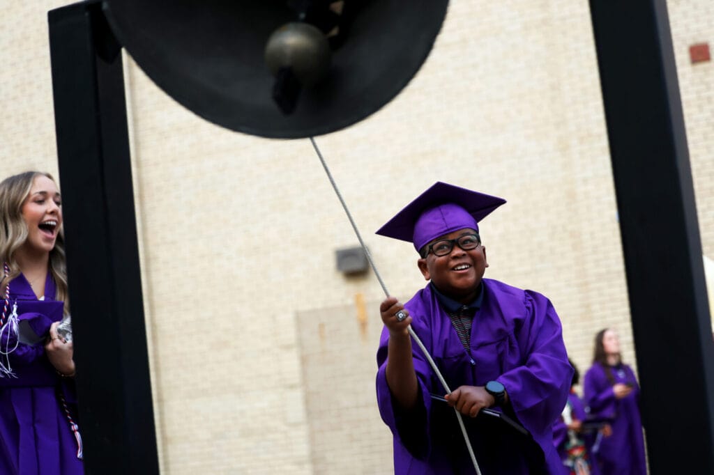 A young man wearing a cap and gown smiles while ringing the victory bell at Bracewell stadium following the commencement ceremony for the Burlington High School class of 2025.