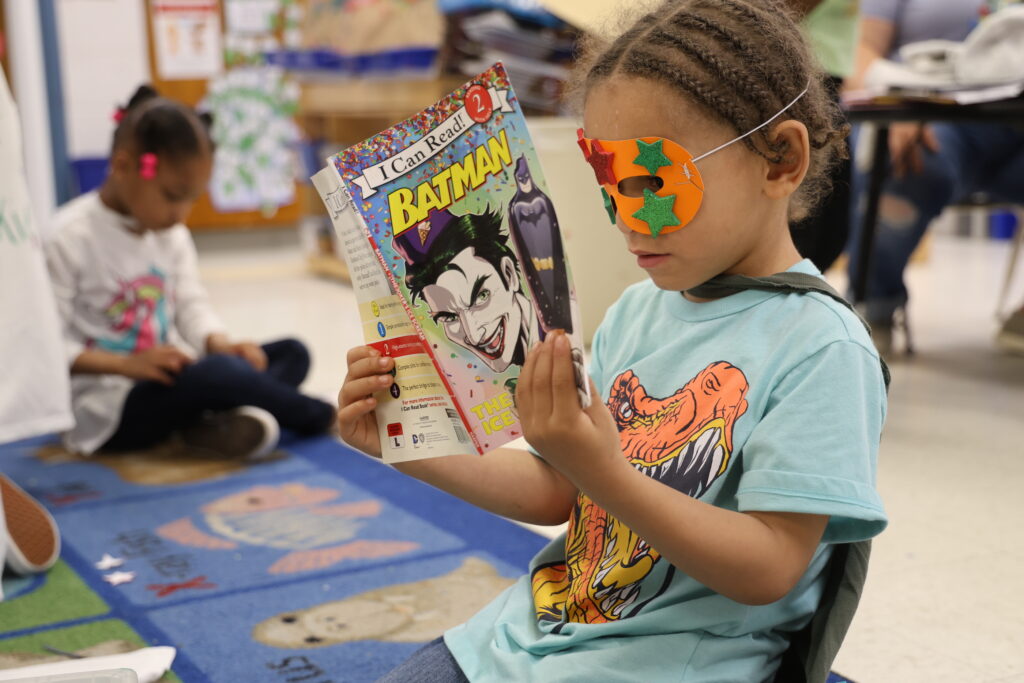 A 4-year-old preschool student wearing a mask and cape reads a "Batman" book during Superhero Day at Corse Early Childhood Center.