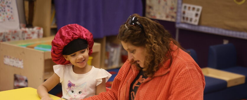 A girl and her preschool teacher sit at a table reviewing a worksheet.