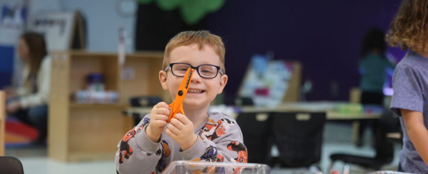 A boy with red hair and glasses in the 3-year-old preschool program holds up an orange pair of safety scissors