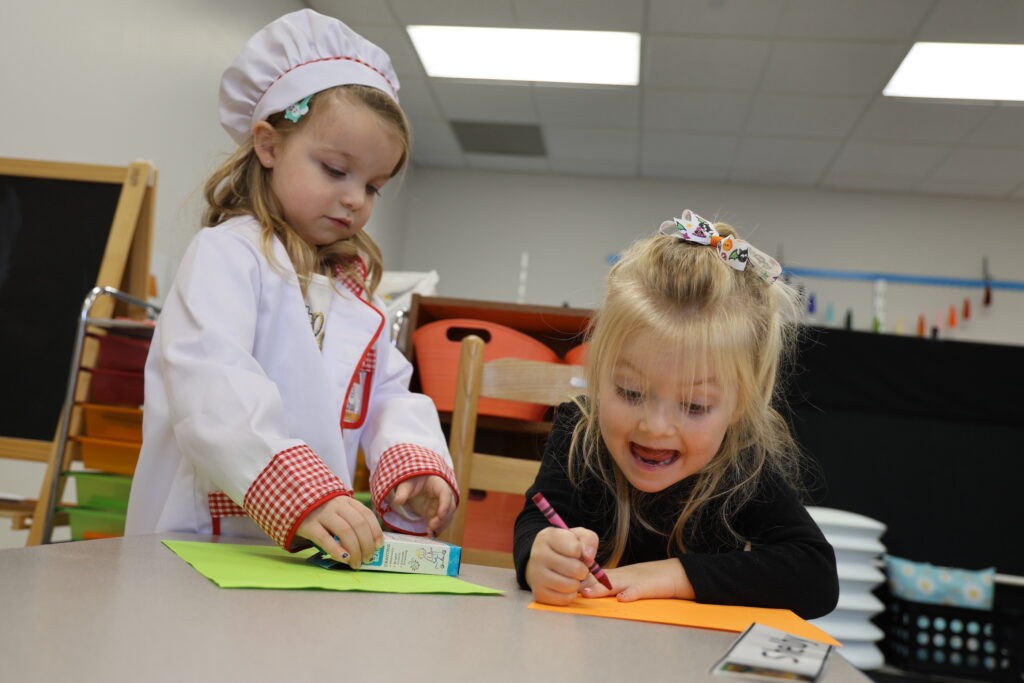 A 3-year-old girl dresses like a chef while getting a crayon out of a box while another 3-year-old girl smiles as she draws the letter "S." 