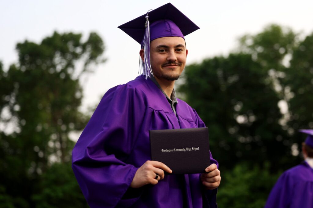 A graduate wearing a purple cap and gown smiles while holding up his diploma during Burlington High School's 2025 commencement ceremony at Bracewell Stadium.