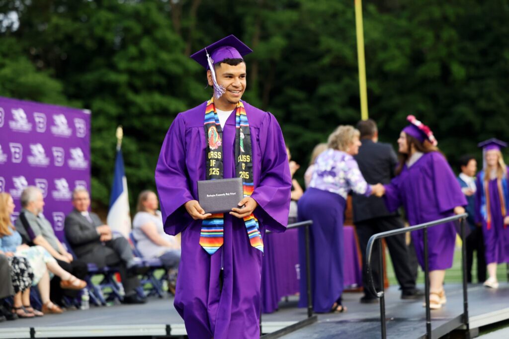 A graduate wearing a purple cap and gown smiles while holding up his diploma during Burlington High School's 2025 commencement ceremony at Bracewell Stadium.
