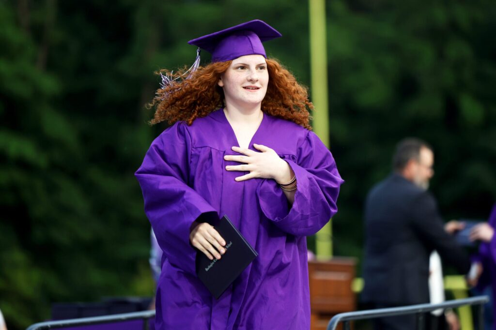 A graduate wearing a purple cap and gown smiles while holding her diploma and returning to her seat during Burlington High School's 2025 commencement ceremony at Bracewell Stadium.