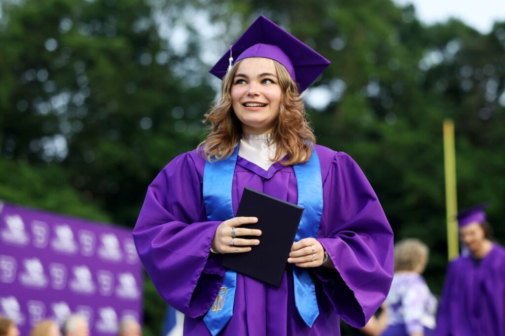 A graduate wearing a purple cap and gown smiles while holding up her diploma during Burlington High School's 2025 commencement ceremony at Bracewell Stadium.