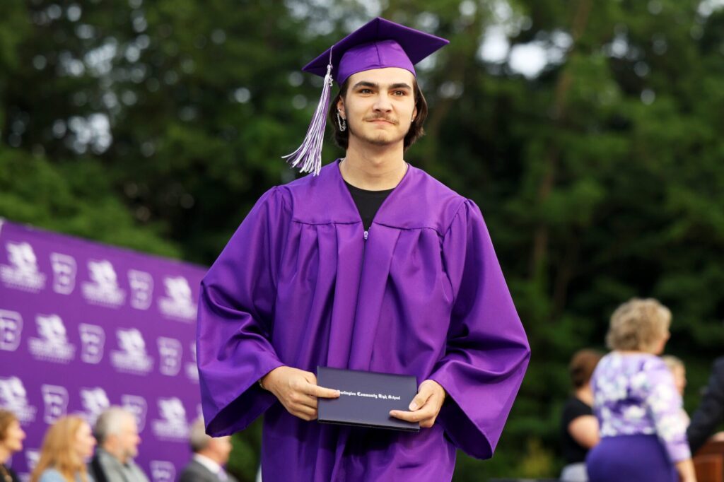 A graduate wearing a purple cap and gown smiles while holding up his diploma during Burlington High School's 2025 commencement ceremony at Bracewell Stadium.