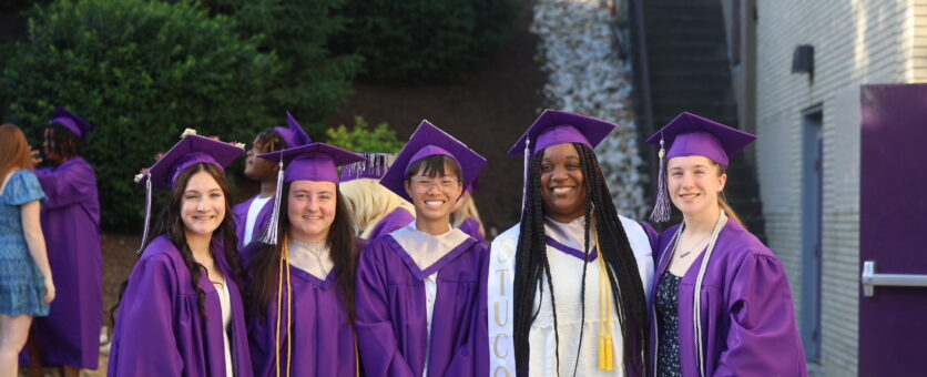 Five girls wearing their caps and gowns stand side by side for a photo ahead of their graduation ceremony.