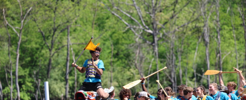 A dragon boat glides through the water while its student passengers cheer and the flag catcher holds up the flag.