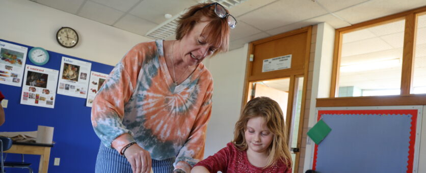 A student and her teacher stand alongside each other at a table with crumpled leaves, paper and glue