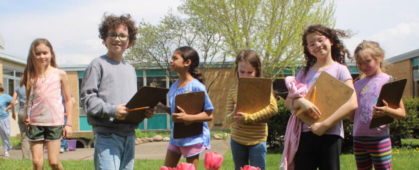 Six students holding clipboards stand outside behind a row of tulips