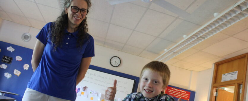 A boys gives two thumbs up while standing beside his teacher