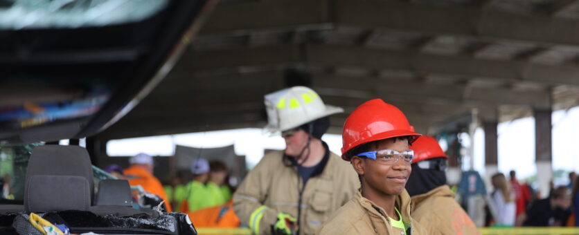 A middle school boy smiles while wearing firefighter gear and safety glasses during an outdoor event featuring an extraction expo put on by the fire department