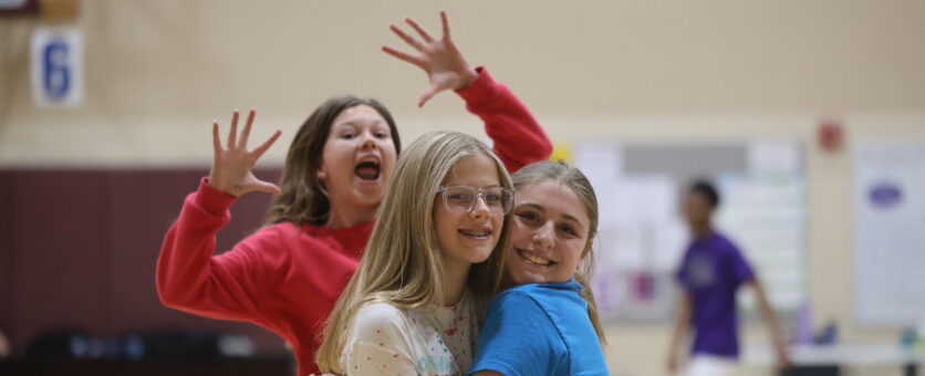 Two middle school girls smile while hugging as a third jumps up behind them in the background