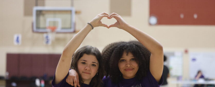 Two middle school girls wearing Burlington Grayhound t-shirts stand next to each other while forming a heart with their hands.